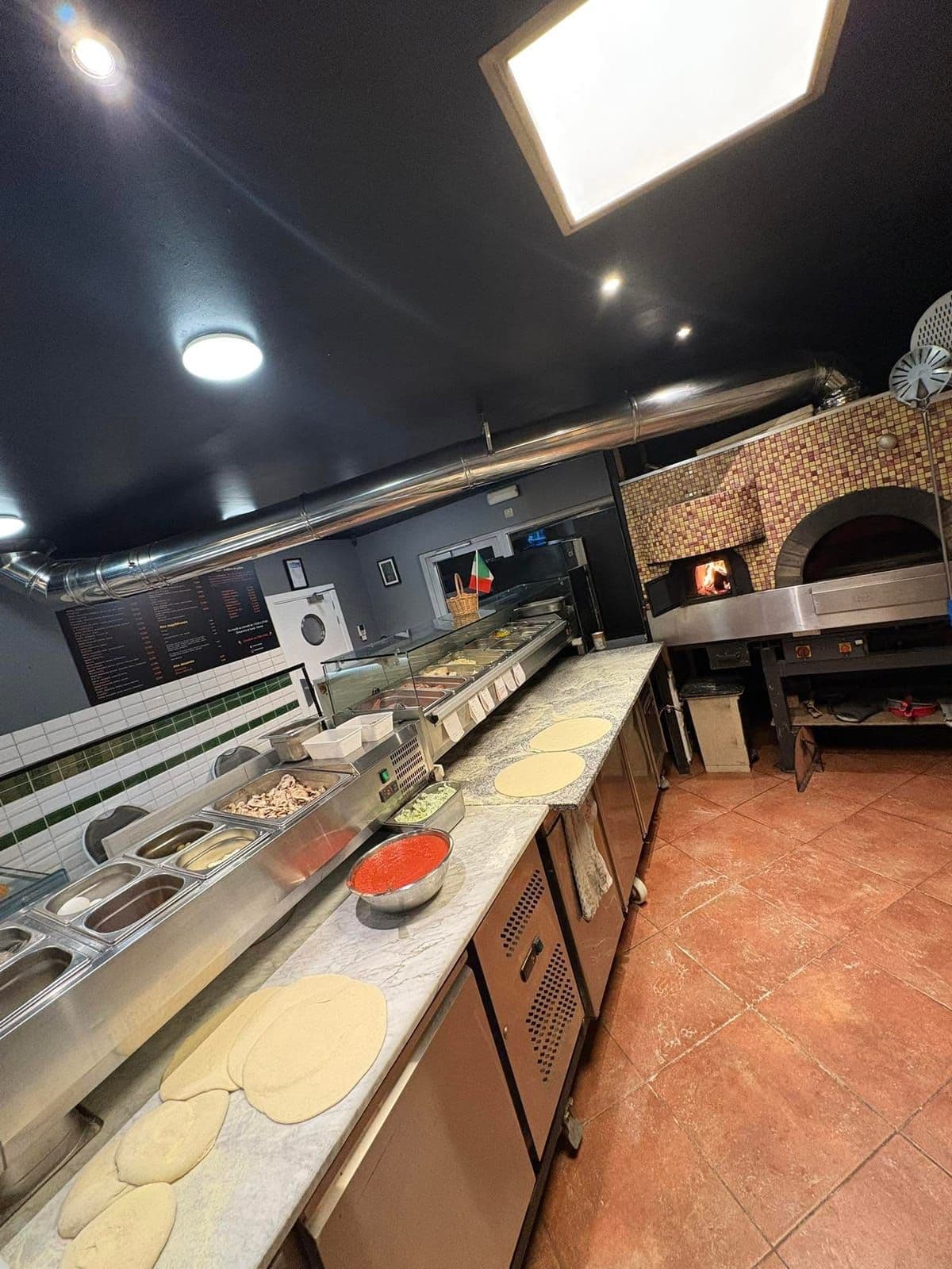 La cuisine de la pizzeria vue depuis l'entrée : le comptoir des garnitures, les pâtons, et le four en mosaïque au fond