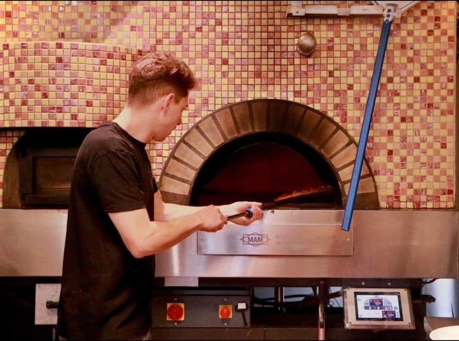 Un pizzaiolo en t-shirt noir enfourne une pizza à la pelle dans le four en mosaïque