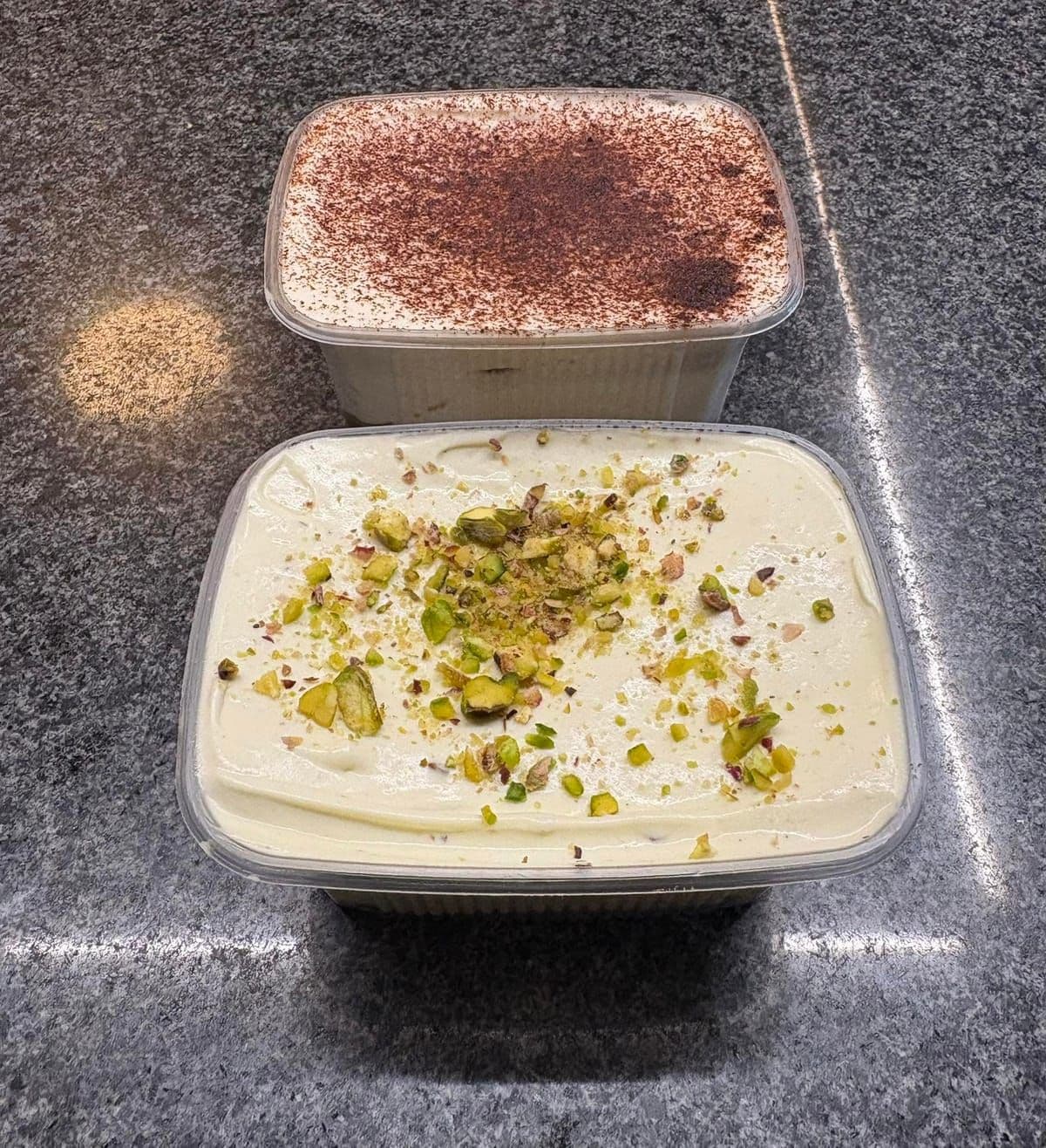 Deux tiramisus en barquette, l'un saupoudré de cacao, l'autre à la pistache, sur le comptoir en granit