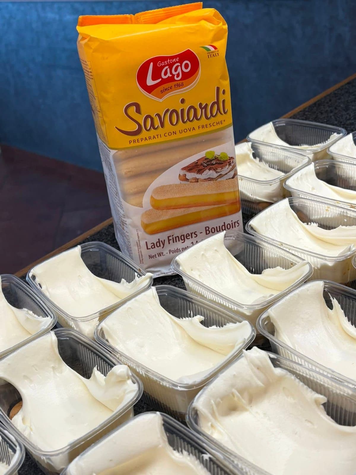 Des barquettes de tiramisu en cours de montage, à côté d'un paquet de biscuits à la cuillère